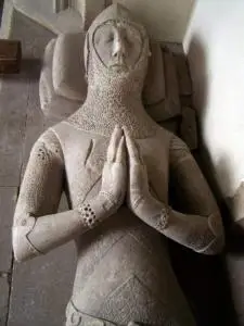 Medieval stone effigy of a knight in armor lying on his back, hands pressed together in prayer, detailed chainmail and facial features visible, set on a tomb in what appears to be a church or cathedral.
