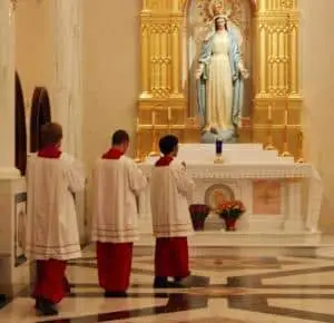 Three altar servers in white and red robes stand facing a statue of the Virgin Mary above an altar, with flowers and candles, honoring Mary's journey of faith in the serene church setting.