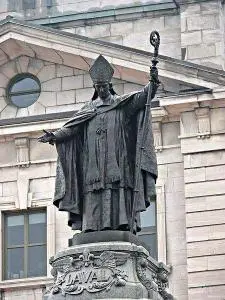 A bronze statue of a bishop holding a staff, wearing traditional religious robes and a mitre hat, stands on a stone pedestal inscribed with LAVAL in front of a stone building with arched windows.