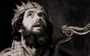 A bearded man wearing a crown and robes looks upward with an expressive face, holding a harp beside him. The image is in dark, dramatic lighting, giving it a classical, historical feel.