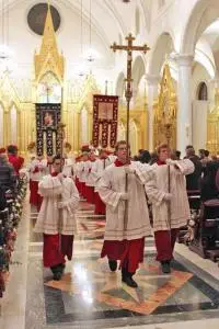 A church procession features altar servers in white and red robes carrying a cross and religious banners down the aisle, with congregants seated on either side in a grand, ornate church with golden decor.