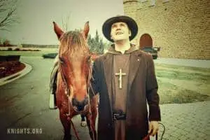 A man dressed in religious clothing with a large cross necklace stands next to a brown horse outside a stone building, looking up and smiling. The scene appears vintage or filtered.