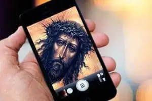 A hand holds a smartphone displaying an image of a man with long hair, a beard, and a crown of thorns, resembling traditional depictions of Jesus, as if taking a photo of a painting or icon.