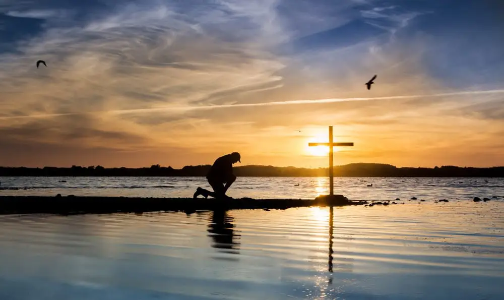 Silhouette of man kneeling in front of cross near body of water at sunset