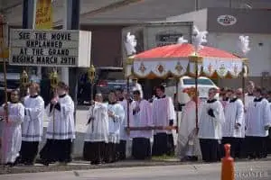 A Catholic procession with priests and altar servers in white and black robes carries a canopy along a street. A marquee in the background advertises the movie Unplanned at The Grand, beginning March 29th.