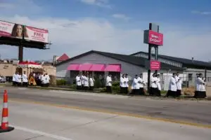 A religious procession of people in white and black robes walks along a sidewalk near a main road, passing by storefronts and a pink sign. Some participants walk under a decorated canopy.