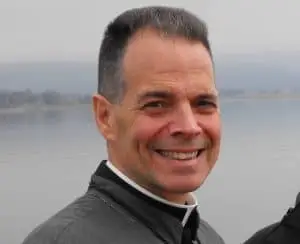 A man with short dark hair, wearing a clerical collar and a grey jacket, smiles at the camera. A body of water and a misty landscape are in the background.