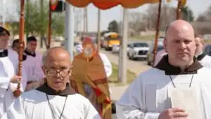 A group of clergy in white robes join an outdoor religious procession devoted to prayer and fraternal life in the spirit of St. Francis of Assisi. A canopy is held overhead, while a monstrance gleams and cars and trees line the street.