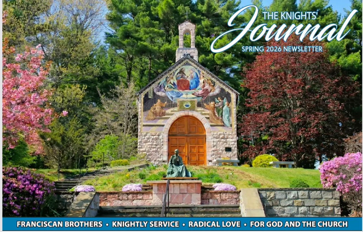A small stone chapel with a colorful mural above its wooden door sits surrounded by lush green trees, pink blossoming flowers, and neatly trimmed bushes. A bronze statue graces the steps in front, featured in the Knights Journal Spring 2026 Newsletter.