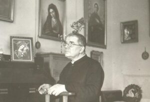 An elderly man in glasses and clerical clothing sits indoors, looking upward. Religious paintings, including portraits of saints and the Virgin Mary, adorn the wall behind him.